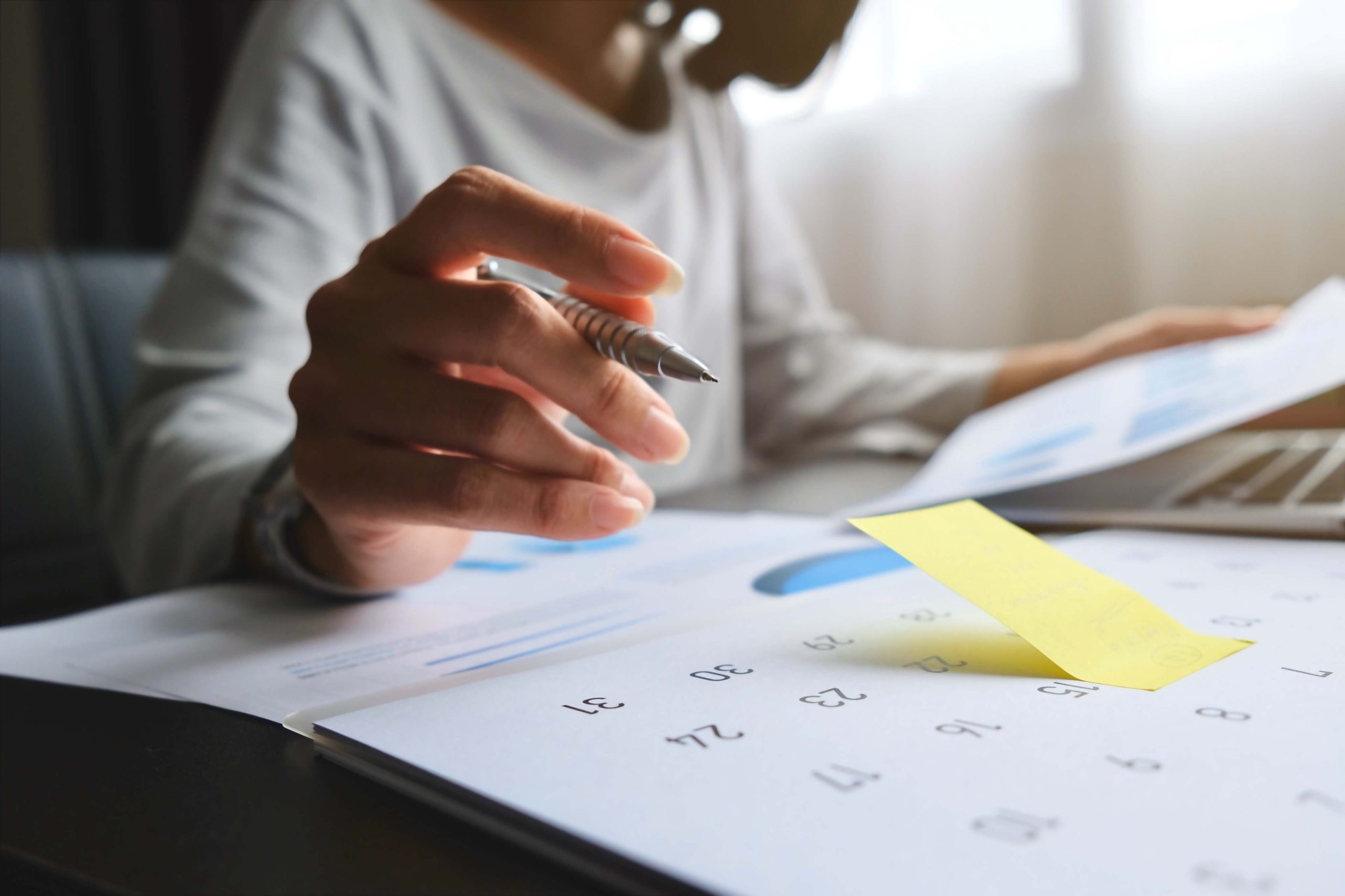 Woman working at desk with large calendar in front of her and pen in hand.